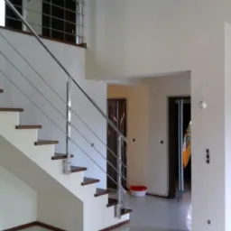 Wykończone wnętrze domu z nowoczesnymi schodami z drewnianymi stopniami i metalowymi poręczami, kremowe ściany, widoczne drzwi i fragment okna na górze.